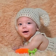 Orion is registered to the contest to win money with this photo: baby, child, knitted_hat, knitted_outfit, smile, happy, toy, orange_toy, green_toy, fluffy_blanket, beige_blanket, cute, portrait, infant, lying_down, indoor, closeup, cozy, warm_colors, button_clothing