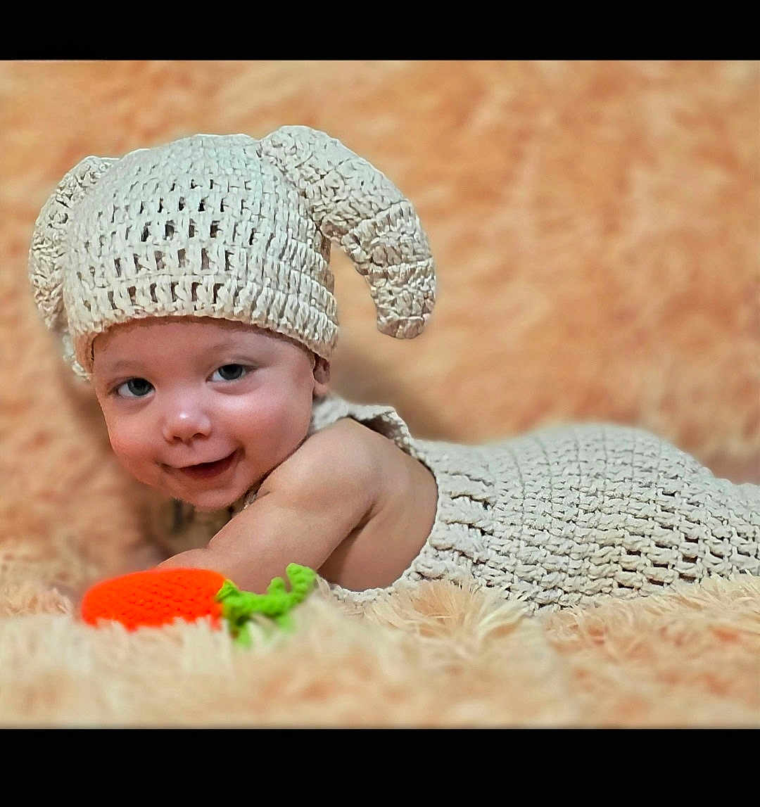 Orion joined the competition — help win amazing prizes! baby, infant, smile, knitted_hat, knitted_outfit, toy, carrot, fluffy_background, cute, lying_down, child, playful, soft_texture, orange, peach_color, happy, portrait, indoors, headwear, hand