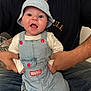 Elijah is registered to the contest to win money with this photo: baby, child, denim_overalls, bucket_hat, marvel, drool, happy, smiling, person, adult_hands, tattoo, couch, cushion, indoor, casual_clothing, jeans, black_tshirt, seated, closeup, portrait