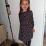 child, girl, smile, dress, floral_pattern, pigtails, sneakers, black_shoes, floor_lamp, wooden_floor, indoor, white_wall, furniture, power_outlet, cables, standing, happy, portrait, casual_clothing, home