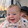 baby, smile, blue_eyes, infant, person, face, clothing, cap, hand, outdoor, sunset, background_blur, happy, cute, young_child, portrait, holding, striped_overall, patterned_shirt, skin
