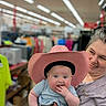 Anastasia is registered to the contest to win money with this photo: baby, woman, cowboy_hat, pink_hat, tattoo, clothing, store, shopping, indoor, smile, child, person, holding, hand, cute, blue_eyes, short_sleeve, casual, colorful, background