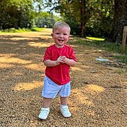 Colton is registered to the contest to win money with this photo: child, toddler, smiling, standing, red_shirt, blue_shorts, white_shoes, outdoor, gravel_path, sunlight, trees, nature, daylight, happy, young_child, portrait, casual_clothing, summer, person, cute