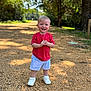 child, toddler, smiling, standing, red_shirt, blue_shorts, white_shoes, outdoor, gravel_path, sunlight, trees, nature, daylight, happy, young_child, portrait, casual_clothing, summer, person, cute