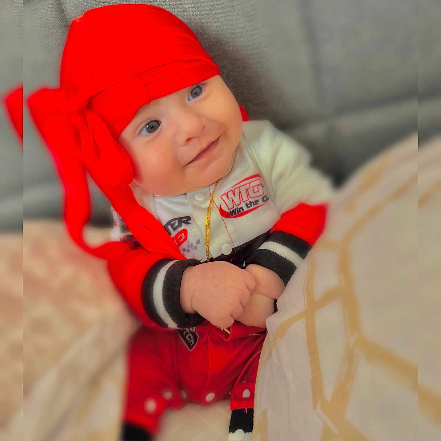 Thiago joined the competition — help win amazing prizes! baby, child, couch, cozy, cushion, cute, face, fashion, gold_chain, hands, happy, indoor, infant, person, portrait, red_clothing, red_headwrap, sitting, smile, white_clothing