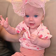 Dorothea is registered to the contest to win money with this photo: baby, blue_eyes, bow, child, couch, cute, face, hand, headband, indoor, infant, patterned_shirt, person, pink_clothing, pink_pants, playful, sitting, smiling, soft_lighting, young_child