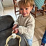 toddler, child, playing, tambourine, indoor, kitchen, sweater, wooden_chair, couch, floor_tiles, smiling, blonde_hair, toy, person, happy, casual_clothing, furniture, home, cute, curly_hair