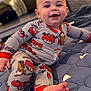 baby, child, smiling, pajamas, clothing, foot, face, indoor, quilt, blanket, toy_car_pattern, happy, cute, toddler, person, sitting, home, living_room, infant, soft_lighting