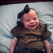 Mia is registered to the contest to win money with this photo: baby, smiling, infant, headband, bow, clothing, pillow, bed, wooden_headboard, cute, happy, portrait, lying_down, child, toddler, apparel, fabric, face, skin, indoors