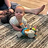 baby, child, plush_toy, rug, floor, wooden_floor, person, adult, legs, barefoot, casual, indoor, home, carpet, toy, colorful, crawling, cute, infant, relaxed