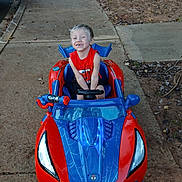 Grady is registered to the contest to win money with this photo: child, smiling, toy_car, red, blue, sidewalk, outdoor, happy, person, play, vehicle, spiderman_theme, concrete, leaves, casual_clothing, blond_hair, daylight, fun, small_car, kid