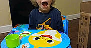 Grady is registered to the contest to win money with this photo: barefoot, blond_hair, blue_chair, cardboard_box, cartoon_characters, casual_clothing, child, colorful, curly_hair, expression, furniture, green_cup, indoor, kids_table, person, room, surprised, table, wooden_floor, young_child