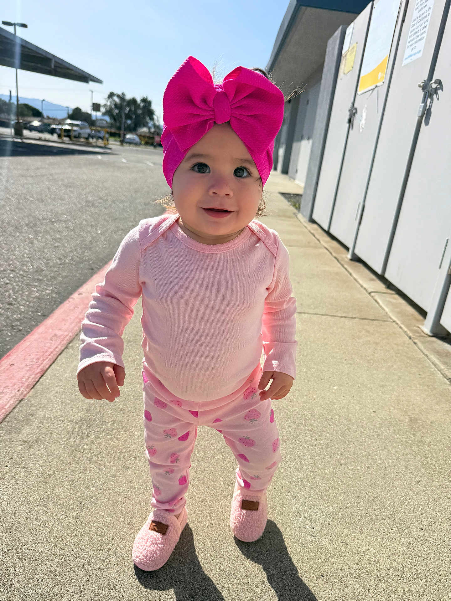 Blake is registered to the contest to win money with this photo: toddler, child, pink_bow, pajamas, outdoor, sidewalk, sunlight, happy, smile, footwear, clothing, person, cute, young_child, daytime, urban, shadow, headwear, walking, playful