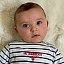 baby, infant, face, blue_eyes, striped_shirt, text_on_clothing, heart, lying_down, blanket, soft_texture, closeup, portrait, cute, child, person, apparel, casual_clothing, indoor, expression, young