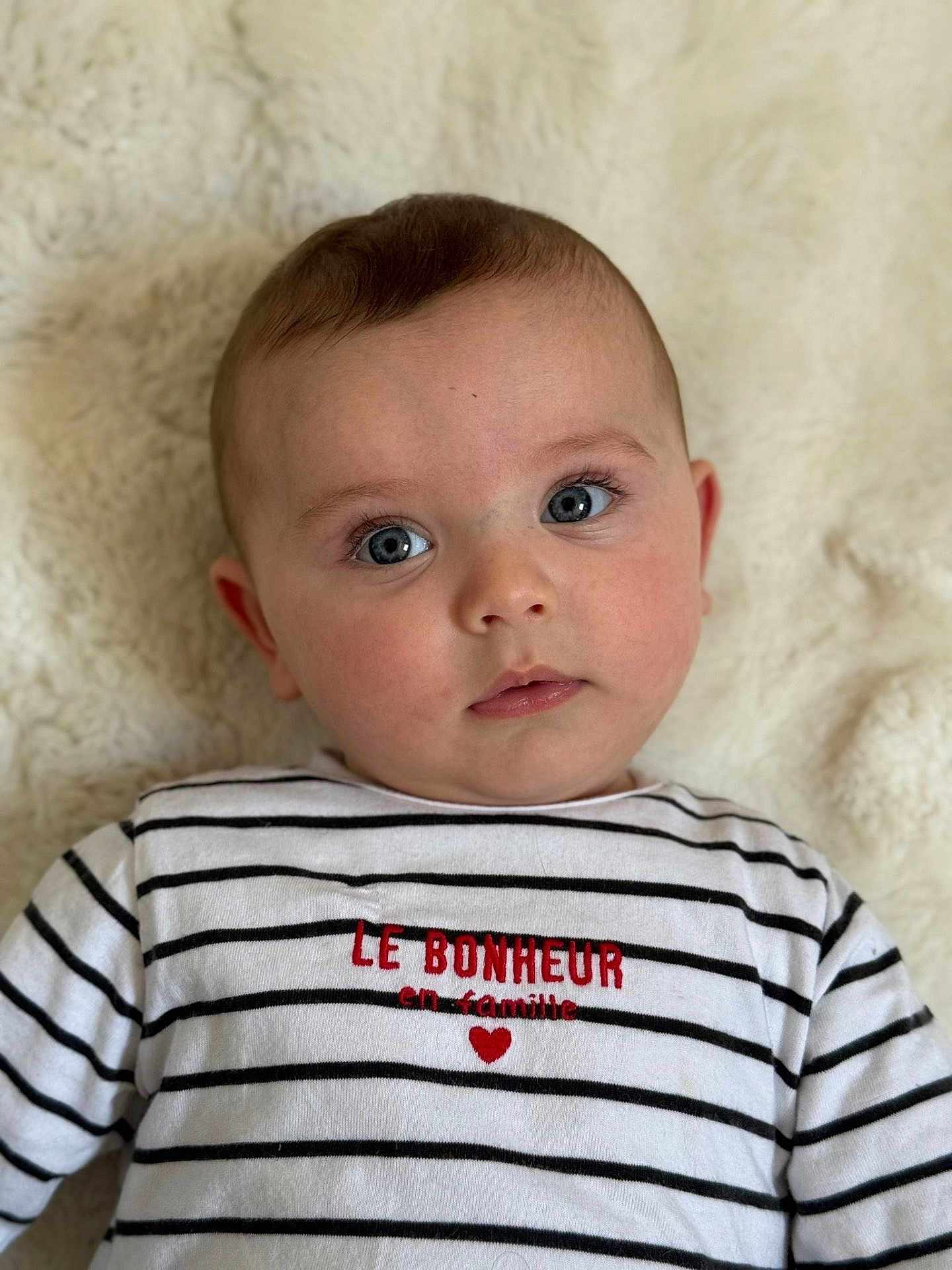 Milann a rejoint le concours — aidez-le/la à gagner de superbes lots ! baby, infant, face, blue_eyes, striped_shirt, text_on_clothing, heart, lying_down, blanket, soft_texture, closeup, portrait, cute, child, person, apparel, casual_clothing, indoor, expression, young
