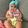 baby, infant, smile, headband, bow, pink_bib, text_on_bib, yellow_outfit, patterned_clothing, couch, gray_couch, seated, chubby_arms, chubby_legs, barefoot, happy, indoors, portrait, cute, child