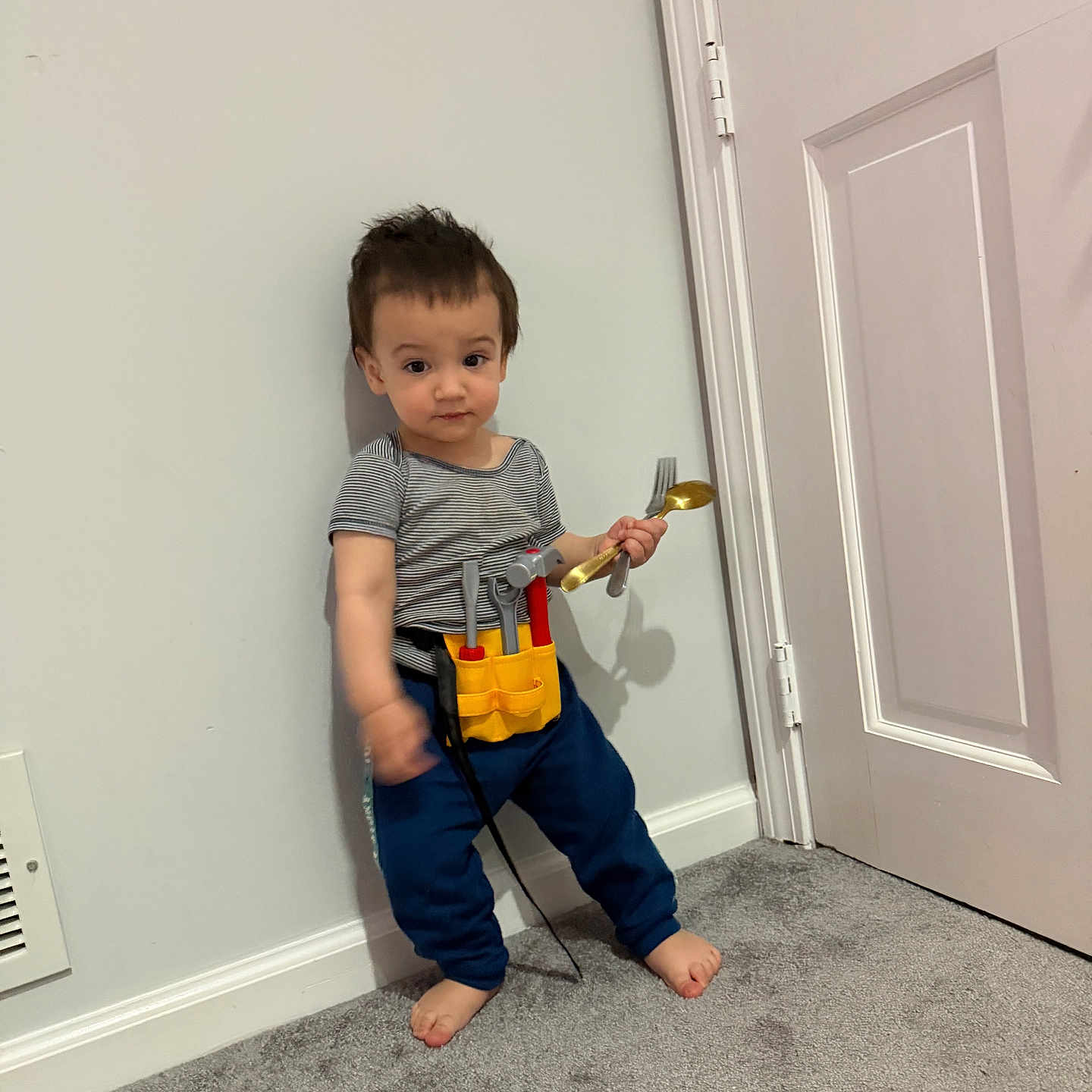 Khalid is registered to the contest to win money with this photo: child, toddler, boy, portrait, indoor, standing, barefoot, tool_belt, toy_tools, spoon, door, wall, carpet, home, playtime, striped_shirt, blue_pants, cute, serious_expression, handyman