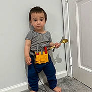 Khalid is registered to the contest to win money with this photo: barefoot, blue_pants, boy, carpet, child, door, fork, full_body, home, indoors, playtime, portrait, serious_expression, spoon, standing, striped_shirt, toddler, tool_belt, toy_tools, wall
