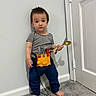 barefoot, blue_pants, boy, carpet, child, door, fork, full_body, home, indoors, playtime, portrait, serious_expression, spoon, standing, striped_shirt, toddler, tool_belt, toy_tools, wall