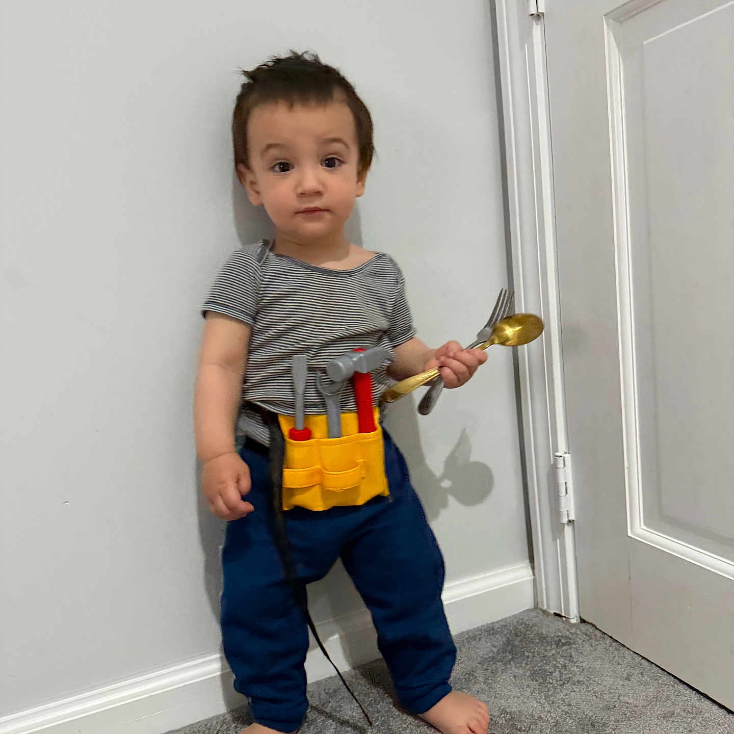 Khalid is registered to the contest to win money with this photo: barefoot, blue_pants, boy, carpet, child, door, fork, full_body, home, indoors, playtime, portrait, serious_expression, spoon, standing, striped_shirt, toddler, tool_belt, toy_tools, wall