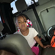Naiely is registered to the contest to win money with this photo: child, car_interior, seat, braids, beads, pink_skirt, white_shirt, seatbelt, backseat, window, playful, pouting, person, vehicle, headrest, daylight, casual_clothing, young_girl, funny_face, colorful_hair_accessories