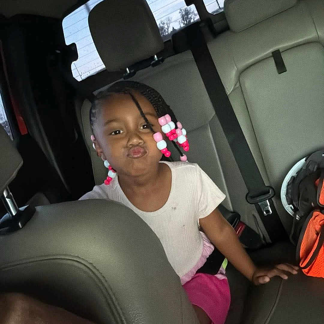 Naiely is registered to the contest to win money with this photo: child, car_interior, seat, braids, beads, pink_skirt, white_shirt, seatbelt, backseat, window, playful, pouting, person, vehicle, headrest, daylight, casual_clothing, young_girl, funny_face, colorful_hair_accessories