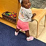 child, toddler, girl, smiling, portrait, standing, bow, hair_accessory, curly_hair, pink_shorts, pink_shoes, sneakers, shirt, toy_basket, colorful_toys, chair, wooden_furniture, carpet, indoor, playful