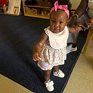 Bradlee is registered to the contest to win money with this photo: toddler, child, baby, standing, smiling, pink_bow, dress, floral_clothing, shoes, socks, classroom, indoor, carpet, wooden_shelves, toys, adult, parent, portrait, full_body, happy