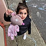 child, girl, plush_toy, mouse_puppet, chalk_drawings, sidewalk, outdoor, black_vest, white_sneakers, playtime, happy, cute, toy, pavement, casual_clothing, daylight, young_child, smiling, looking_up, hand_held_object