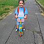 child, scooter, smile, outdoor, backpack, pavement, grass, shorts, sneakers, happy, person, topknot, play, daytime, urban, path, casual_clothing, fun, young, standing