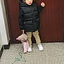 child, indoor, door, black_vest, beige_pants, white_shoes, stuffed_toy, mouse_toy, pink_sweater, tile_floor, string, standing, person, casual_clothing, young_child, holding, toy, footwear, brown_door, wall