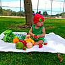 Valentina is registered to the contest to win money with this photo: baby, blanket, child, colorful, curious, fence, fruit, grass, green_outfit, lettuce, nature, outdoor, park, picnic, playful, red_hat, summer, sunny, toy, tree