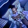 child, baby, money, dollar_bills, striped_cushion, blanket, indoor, hand, curious, laying_down, soft_lighting, blue_tone, cash, infant, clothing, person, home, comfort, relaxing, counting