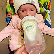 Tessa is registered to the contest to win money with this photo: baby, bottle, feeding, hand, hat, infant, newborn, person, pink_clothing, cushion, cartoon, colorful, lying_down, milk, nursery, cute, soft, indoors, feeding_time, care