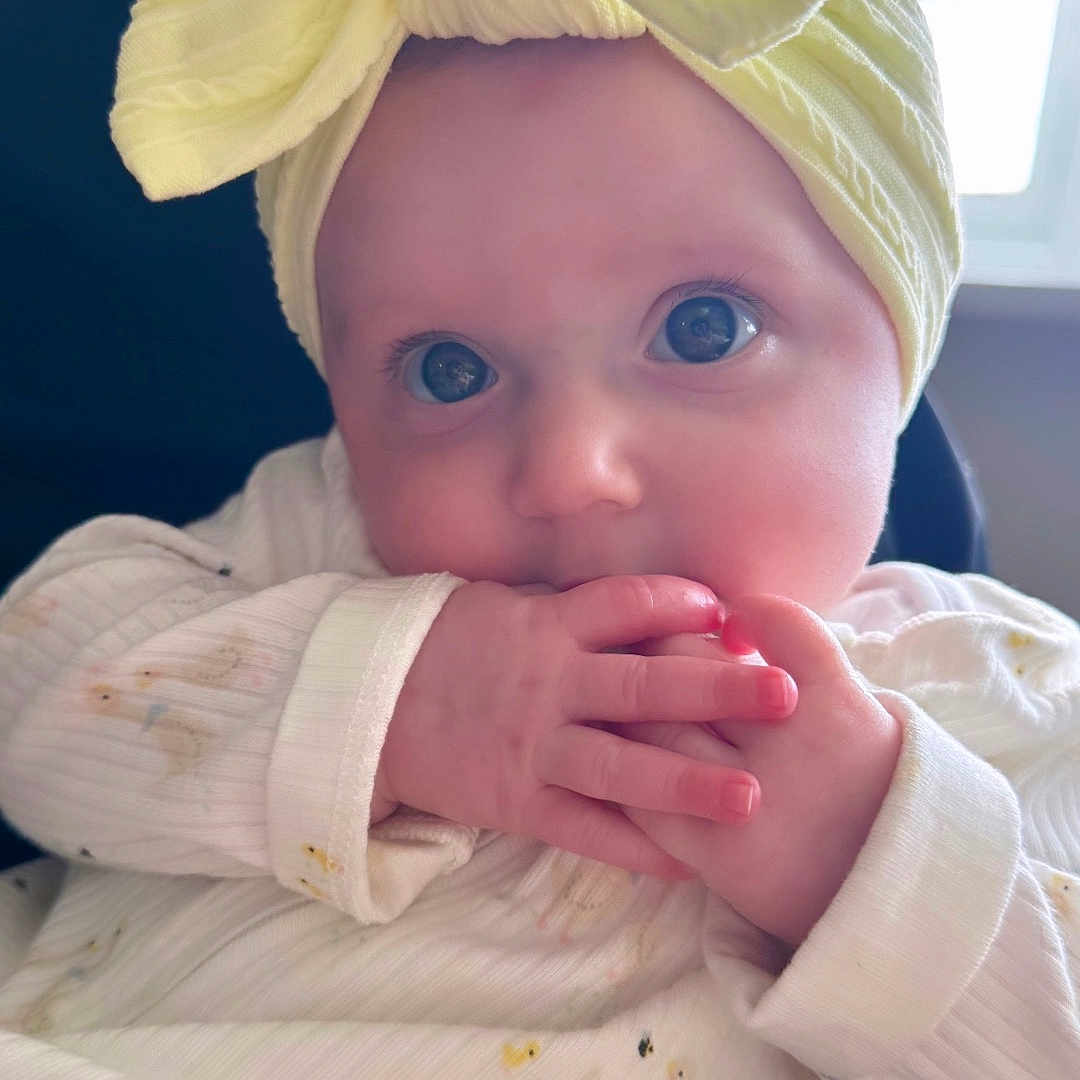 Flo joined the competition — help win amazing prizes! baby, chair, child, close_up, curious, duck_pattern, eyes, face, fingers, hands, headband, indoor, infant, natural_light, portrait, sitting, sucking, white_outfit, window, yellow_bow
