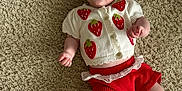 Flo is registered to the contest to win money with this photo: baby, infant, child, smiling, tongue_out, headband, bow, knitted_cardigan, strawberry_pattern, red_shorts, lace_trim, white_socks, carpet, textured_rug, floor, cute, playful, happy, lying_down, indoors