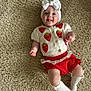 baby, infant, child, smiling, tongue_out, headband, bow, knitted_cardigan, strawberry_pattern, red_shorts, lace_trim, white_socks, carpet, textured_rug, floor, cute, playful, happy, lying_down, indoors