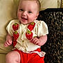 Flo is registered to the contest to win money with this photo: baby, infant, smiling, couch, cushion, knitted_clothing, red_shorts, strawberry_pattern, happy, child, cute, indoors, portrait, sitting, soft_lighting, young_child, cozy, plush, buttoned_sweater, relaxed