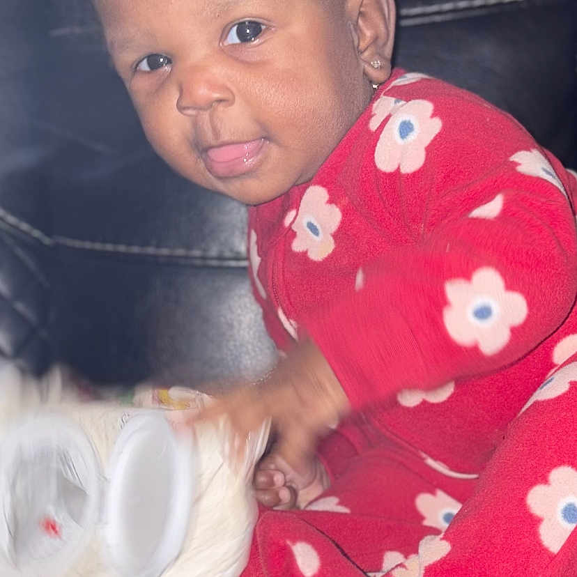 Amirerose is registered to the contest to win money with this photo: toddler, child, red_onesie, floral_pattern, smiling, tongue_out, curly_hair, earrings, black_leather_couch, indoor, playful, holding_object, toilet_paper, baby, person, cute, portrait, sitting, young_child, happy