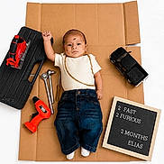 Elias is registered to the contest to win money with this photo: baby, infant, cardboard, toy_tools, toy_car, jeans, white_shirt, sign, milestone, portrait, floor, child, cute, studio, photography, young, person, casual_clothing, indoor, props