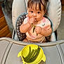 baby, child, high_chair, green_beans, food, eating, bib, pigtails, hair_clips, indoor, kitchen, table, yellow_bowl, person, cute, infant, feeding, portrait, curious, hand