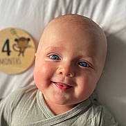 Arthur is registered to the contest to win money with this photo: baby, infant, smiling, blue_eyes, face, head, clothing, gray_outfit, blanket, bed, wooden_plaque, monkey_illustration, milestone, 4_months, portrait, cute, cheeks, happy, lying_down, closeup