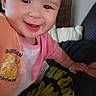 baby, child, smile, face, pink_clothing, orange_clothing, cartoon, indoor, person, cute, happy, closeup, infant, young_child, apparel, hand, couch, blanket, wall, portrait