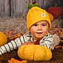 baby, child, hat, yellow_hat, plush_pumpkin, autumn_leaves, brown_rug, striped_shirt, fall_decor, cute, smiling, indoors, portrait, cozy, seasonal, soft_toys, pumpkins, happy, young_child, headwear