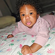 Trui is registered to the contest to win money with this photo: baby, child, curly_hair, pink_clothing, blanket, handprint_pattern, tongue_out, face, portrait, infant, cute, lying_down, expression, indoors, soft_lighting, skin, person, young_child, playful, closeup