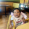 Reign is registered to the contest to win money with this photo: baby, child, high_chair, bib, smiling, happy, restaurant, table, chair, wood, indoor, floor_tiles, window, sunlight, cushion, clothing, person, cute, portrait, infant