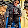 child, toddler, playground, slide, sitting, outdoor, autumn, leaves, jacket, winter_clothing, hat, pants, shoes, smiling, portrait, playground_equipment, chain_link_fence, sunlight, park, play