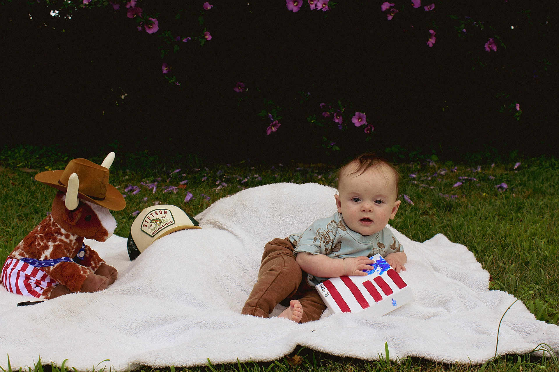 Stetson joined the competition — help win amazing prizes! baby, blanket, grass, outdoor, toy, plush_bull, cowboy_hat, american_flag, craft, child, greenery, purple_flowers, shirt, pants, foot, nature, cute, sitting, grass_field, playtime