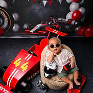 Riyank is registered to the contest to win money with this photo: baby, balloons, beanbag, black, checkered_flags, child, cute, decor, floor, helmet, indoor, playful, portrait, racing, red, smiling, sunglasses, toy_car, toy_motorcycle, white
