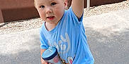 Gemma is registered to the contest to win money with this photo: background_wall, blue_clothing, casual, child, concrete, curious_expression, daylight, footwear, gravel, holding_object, metal_bar, outdoor, person, pink_shoes, playful, short_hair, sippy_cup, standing, toddler, young_child